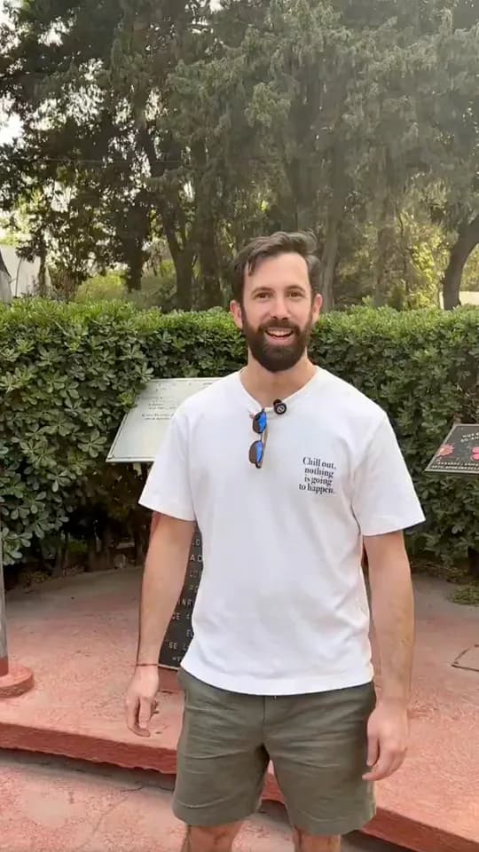 A man in a white T-shirt smiles in front of a garden and a monument in Mexico City