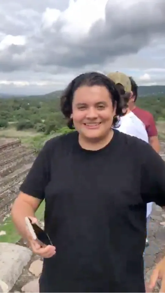 A man smiles holding his phone at the top of a Teotihuacán pyramid under a cloudy sky